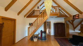 Geräumiges Wohnzimmer mit hohen Holzdecken, Galerie und Kaminofen, strahlt Gemütlichkeit aus.