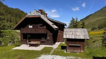Traditionelles Holzhaus im Chalet-Stil mit Balkon und gepflasterter Terrasse in alpiner Landschaft.