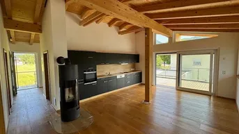 Offener Wohn-Essbereich mit moderner Küche, Kaminofen und sichtbaren Holzbalken an der Decke.