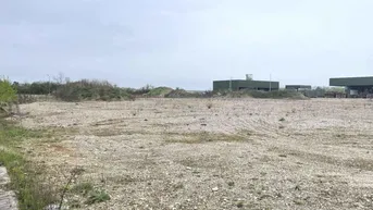 Großes, unbebautes Grundstück mit Kiesboden und spärlicher Vegetation, im Hintergrund sind Industriegebäude sichtbar.