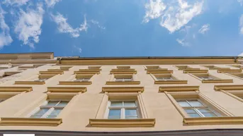 Blick nach oben auf die Fassade eines Altbaus mit vielen Fenstern unter einem blauen Himmel mit Wolken.