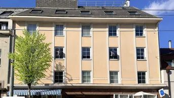 Gepflegte Fassade eines Mehrfamilienhauses mit beiger Putzfassade, Sprossenfenstern und Mansarddach.