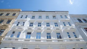Weiße Fassade eines historischen Gebäudes mit vielen Fenstern und dekorativen Elementen unter blauem Himmel.