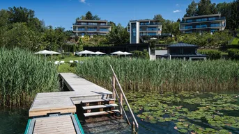 Malerischer Seeblick mit Holzsteg, Schilf und modernen Wohngebäuden am bewaldeten Hang.