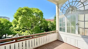 Heller Balkon mit Holzboden, weißem Geländer und Blick ins Grüne unter blauem Himmel.