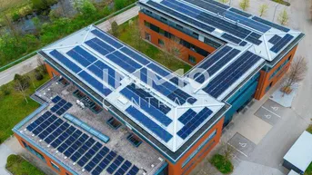 Luftaufnahme eines modernen Bürogebäudes mit zahlreichen Solarmodulen auf dem Dach, umgeben von grünen Feldern und einem Fluss.