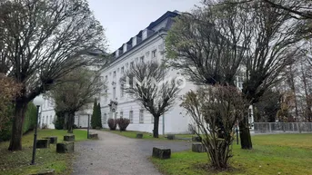 Großes, gut erhaltenes Gebäude mit Mansarddach, umgeben von einem parkähnlichen Garten mit alten Bäumen.