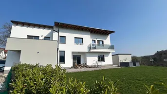 Modernes Einfamilienhaus mit weißer Fassade, Terrasse und Balkon, umgeben von Grünflächen.