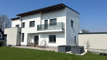 Rückansicht des modernen Hauses mit Terrasse, Balkon und gepflegtem Rasen unter blauem Himmel.