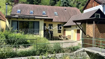 Charmantes Haus mit Satteldach, Dachfenstern und einem kleinen Balkon, umgeben von viel Grün und mit Bergblick.