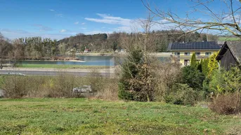 Panoramablick auf einen See mit angrenzender Straße und einem Gebäude mit Solaranlage am Ufer.