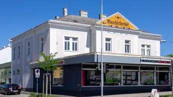 Ansicht des Eckgebäudes mit Geschäftsflächen im Erdgeschoss und Wohnungen darüber, an einer belebten Straße gelegen.
