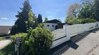 Einfamilienhaus mit weißem Zaun, gepflasterter Auffahrt und üppiger Vegetation unter blauem Himmel.