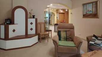 Geräumiges Wohnzimmer mit bequemen Sesseln, einem Kachelofen und Blick in den Flur.