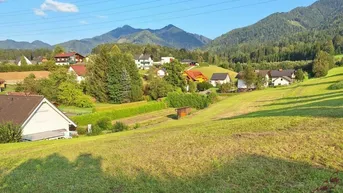 Weitläufige grüne Landschaft mit Häusern, Bergen und blauem Himmel im Hintergrund.