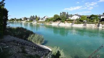 Weitläufiger Blick über den ruhigen See mit Häusern am gegenüberliegenden Ufer unter blauem Himmel.