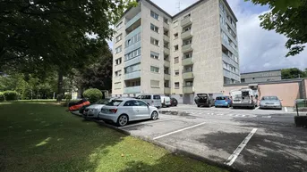 Mehrstöckiges Wohngebäude mit Parkplätzen und Grünflächen in einer städtischen Umgebung.
