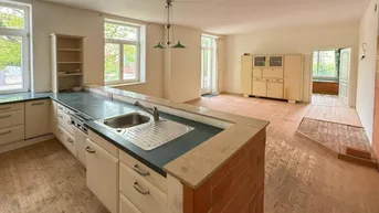 Offener Wohnbereich mit integrierter Küche, Holzdielenboden und Blick ins Grüne durch große Fenster.