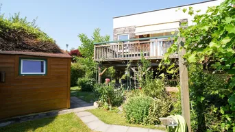 Gepflegter Gartenbereich mit Gartenhaus, Holzterrasse und üppiger Vegetation unter blauem Himmel.