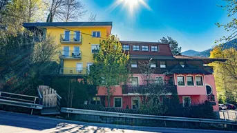 Mehrstöckiges Gebäude mit gelber und rosa Fassade, umgeben von Bäumen unter strahlendem Sonnenschein.