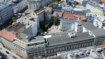 Luftaufnahme eines dicht bebauten Stadtblocks mit verschiedenen Gebäuden, Flachdächern und einem Innenhof mit Parkplätzen.
