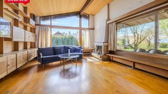 Helles Wohnzimmer mit Holzboden, hohen Decken, Kamin und großen Fenstern mit Blick ins Grüne.