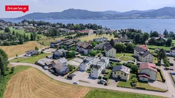 Luftaufnahme einer Wohnsiedlung am Seeufer mit umliegenden Feldern und Bergen im Hintergrund.
