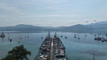 Panoramablick auf einen See mit zahlreichen Segelbooten an einem langen Steg und Bergen im Hintergrund.