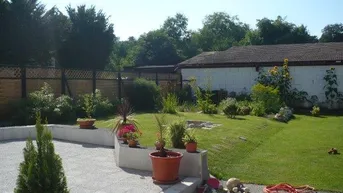 Gepflegter Gartenbereich mit Rasenfläche, befestigtem Weg und blühenden Pflanzen an einem sonnigen Tag.