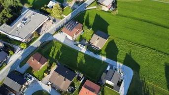 Luftaufnahme einer Wohnsiedlung mit Einfamilienhäusern, grünen Wiesen und Bergen im Hintergrund.
