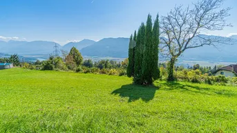 Weitläufige grüne Wiese mit Blick auf die umliegende Berglandschaft und einen See in der Ferne.