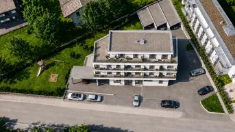 Luftaufnahme eines modernen Mehrfamilienhauses mit Flachdach, Parkplätzen und Grünflächen inklusive Spielplatz.