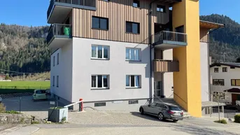 Modernes Mehrfamilienhaus mit Holzfassade, Balkonen und Parkplätzen vor alpiner Kulisse.