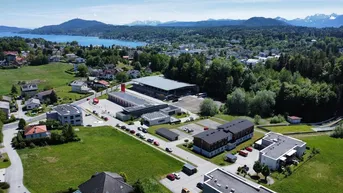 Weitläufige Luftaufnahme einer ländlichen Gegend mit einem See, Bergen im Hintergrund und verschiedenen Gebäuden inmitten grüner Natur.