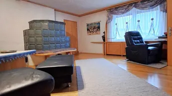 Gemütliches Wohnzimmer mit traditionellem Kachelofen, Holzboden und Sitzgelegenheiten.