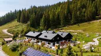 Mehrere moderne Gebäude in alpiner Landschaft mit Photovoltaikanlage und Wald im Hintergrund.