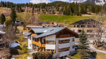 Haus in alpiner Landschaft mit grünen Hügeln und schneebedeckten Bergen im Hintergrund.