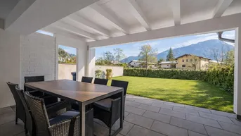 Geräumige überdachte Terrasse mit Esstisch, Blick auf den grünen Garten und die Berge.