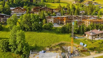Luftaufnahme einer Baustelle in einer alpinen Landschaft mit umliegenden Chalets und grünen Hügeln.