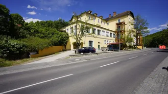 Gelbes Mehrfamilienhaus an einer Straße, umgeben von grünen Bäumen und Hügeln unter blauem Himmel.