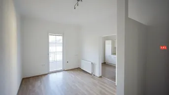 Helles Wohnzimmer mit Laminatboden und Zugang zu einem kleinen Balkon, offen zur Küche.