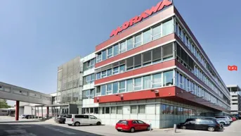 Modernes Bürogebäude mit roter und grauer Fassade, großen Fenstern und angrenzendem Parkplatz unter blauem Himmel.