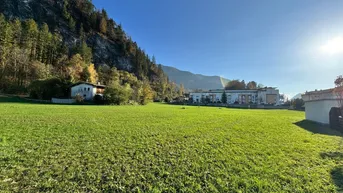 Weitläufige grüne Wiese mit bewaldeten Bergen und Gebäuden im Hintergrund unter strahlend blauem Himmel.