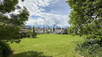Grünes Feld mit Blick auf die Berge und umliegende Häuser unter bewölktem Himmel.