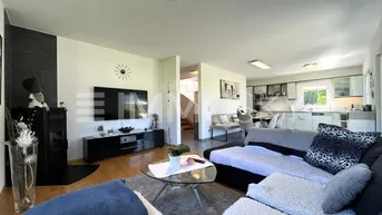 Helles Wohnzimmer mit Kaminofen, Holzboden und offenem Übergang zur Küche.