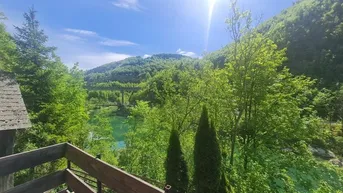 Sonniger Ausblick von einem Balkon auf einen Fluss, eine Brücke und bewaldete Berge.
