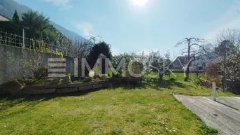 Sonniger Gartenbereich mit Rasenfläche, Holzterrasse und Blick auf die umliegenden Berge.