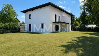 Modernes Einfamilienhaus mit weißer Fassade, großem Rasen und umgebenden Bäumen unter blauem Himmel.
