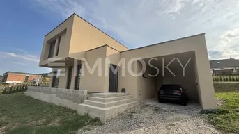 Modernes Einfamilienhaus mit beiger Fassade und integriertem Carport, umgeben von Grünflächen.