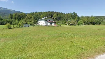 Panoramablick über eine grüne Wiese mit Bergen im Hintergrund und einem Haus in der Ferne.